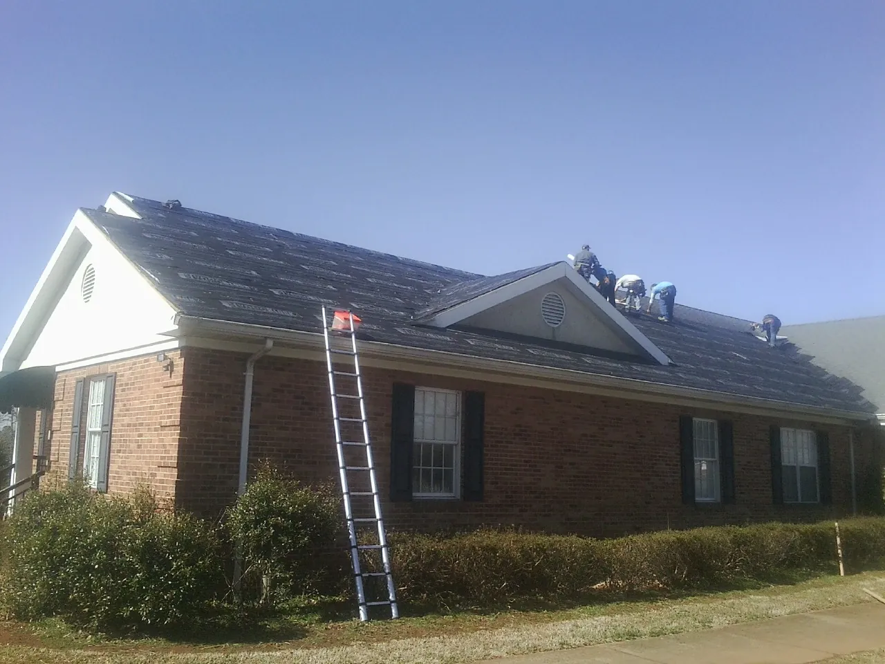 Crew tearing shingles off a brick ranch roof with a ladder set — Emergency Roof Repair work by our crew in Unadilla
